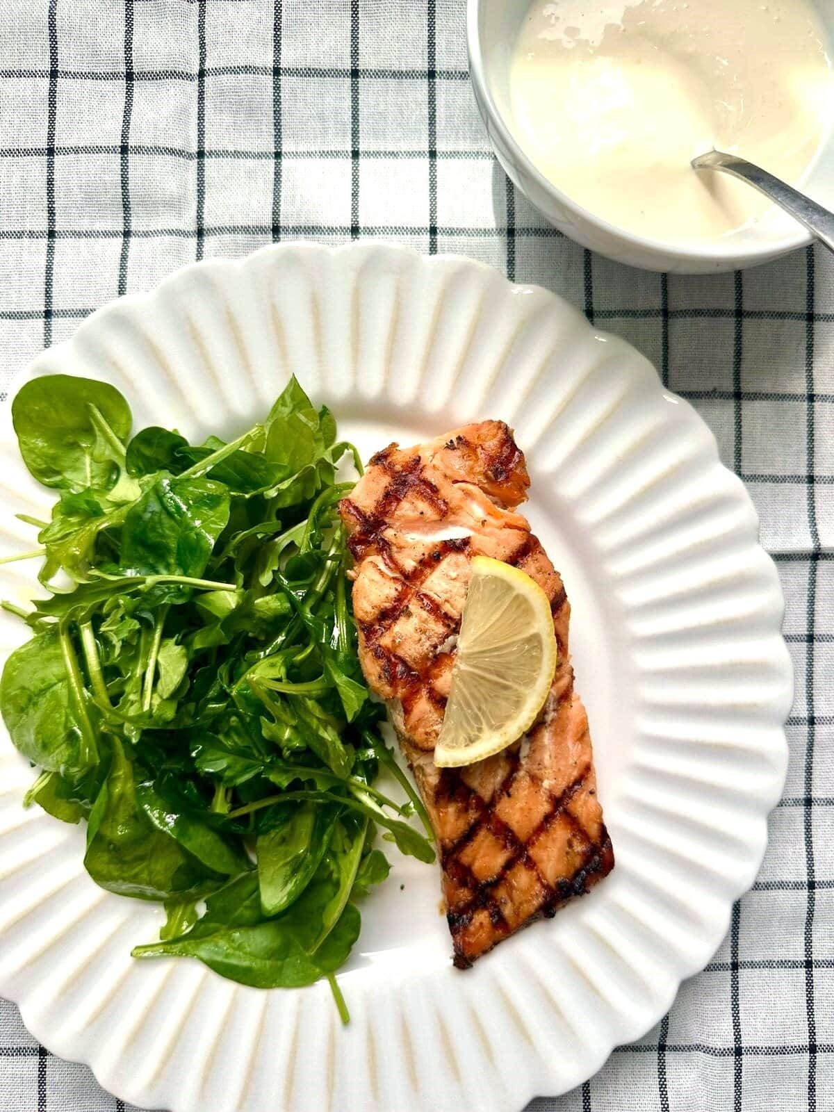 Grilled salmon fillet served with arugula and a slice of fresh lemon with a bowl of lemon aioli set alongside.