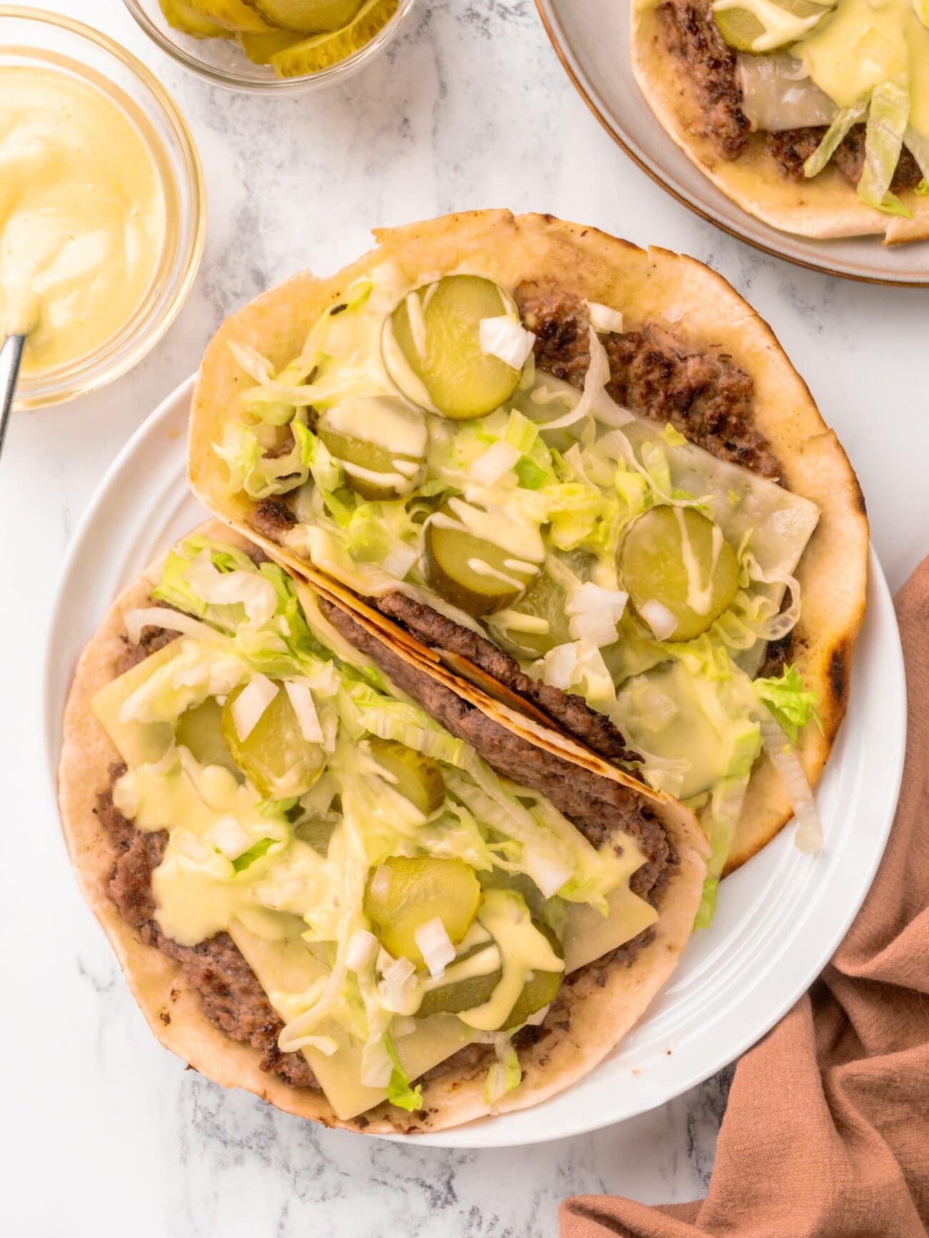 A plate of big mac tacos, topped with ground beef, melted cheese, shredded iceberg lettuce, sliced pickles, diced onion and sauce. With bowls of sauce and sliced pickles set alongside.