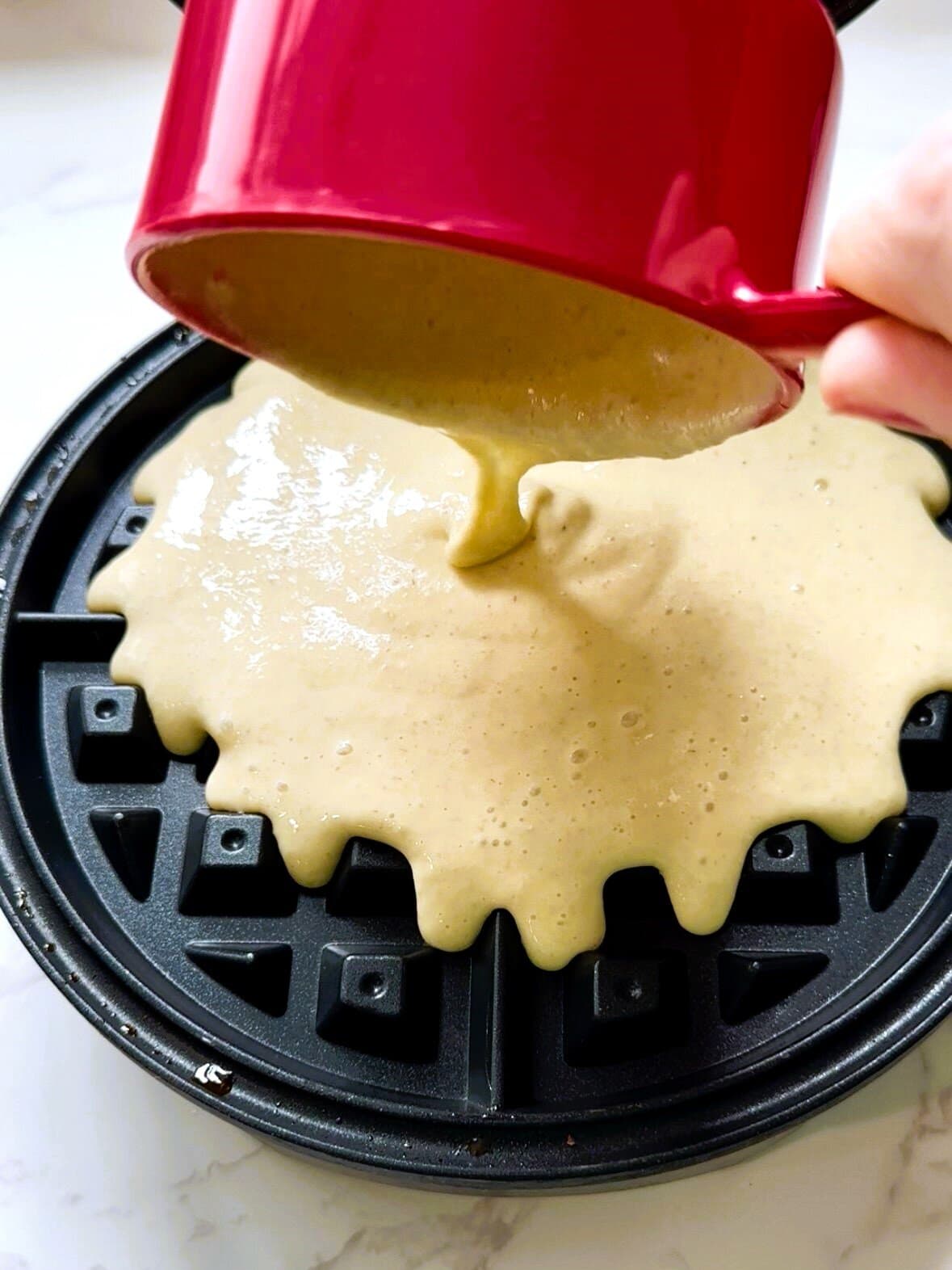 A cup of waffle batter being poured onto a hot waffle iron.