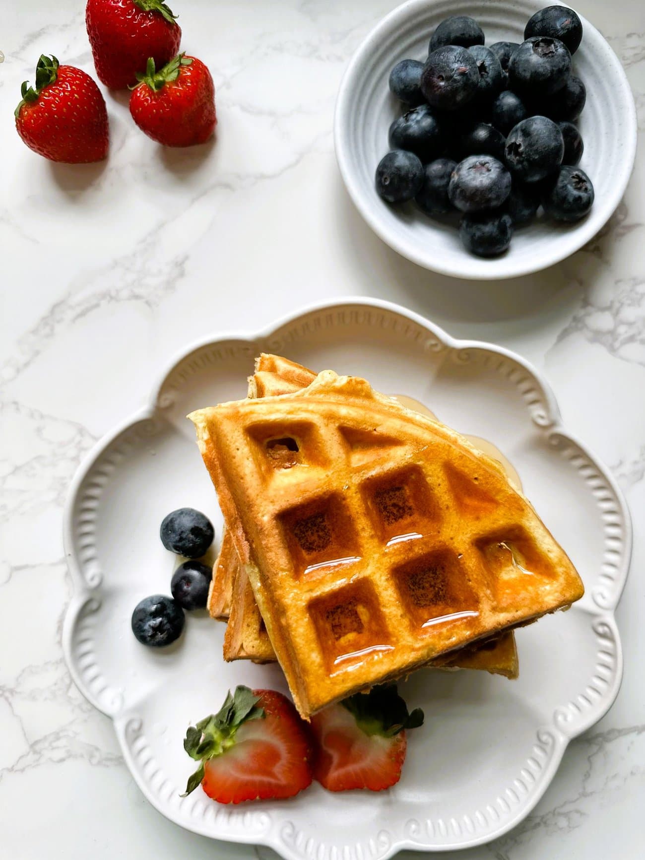 A white plate topped with 3 ingredient high protein waffles topped with strawberries, blueberries and maple syrup, with fresh berries set alongside.