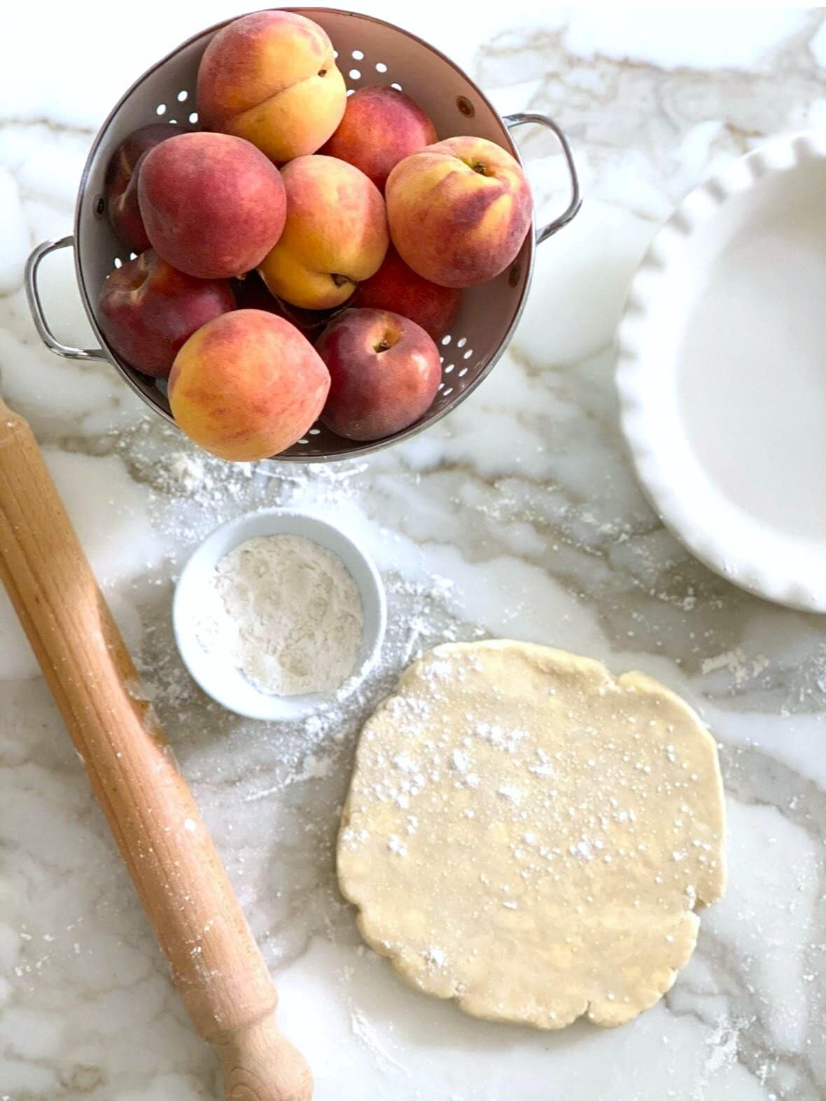 A sheet of pastry dough being rolled out with a rolling pin, with a pie dish and fresh peaches set alongside.