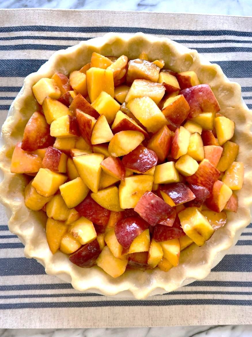 A pie dish covered with a sheet of shortcrust pastry and topped with chopped peaches.