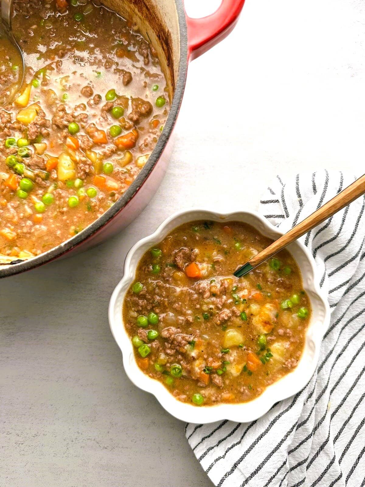 A bowl of hearty shepherd's pie soup garnished with freshly chopped chives, and a pot of soup set alongside.