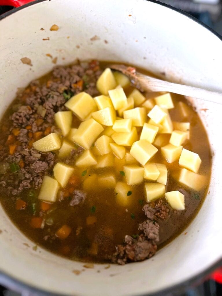A Dutch oven of Shepherd's pie soup with diced potatoes being added.