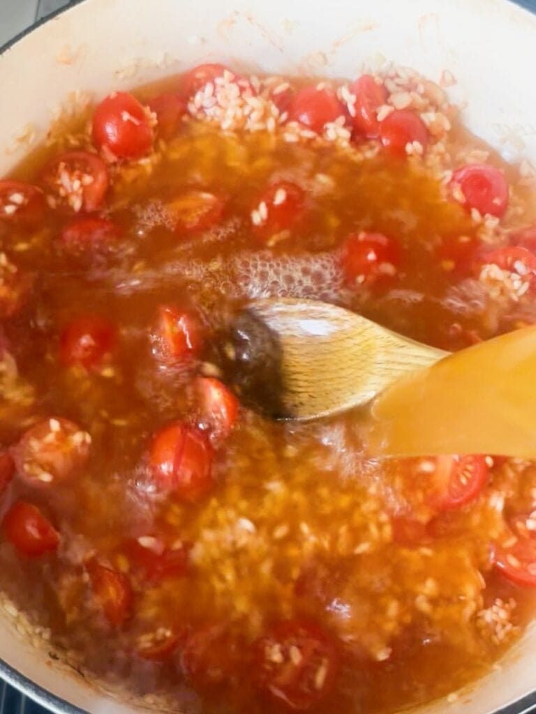 A large skillet with sauteed onion, garlic rice, with added tomato paste and fresh cherry tomato halves, with added vegetable stock.