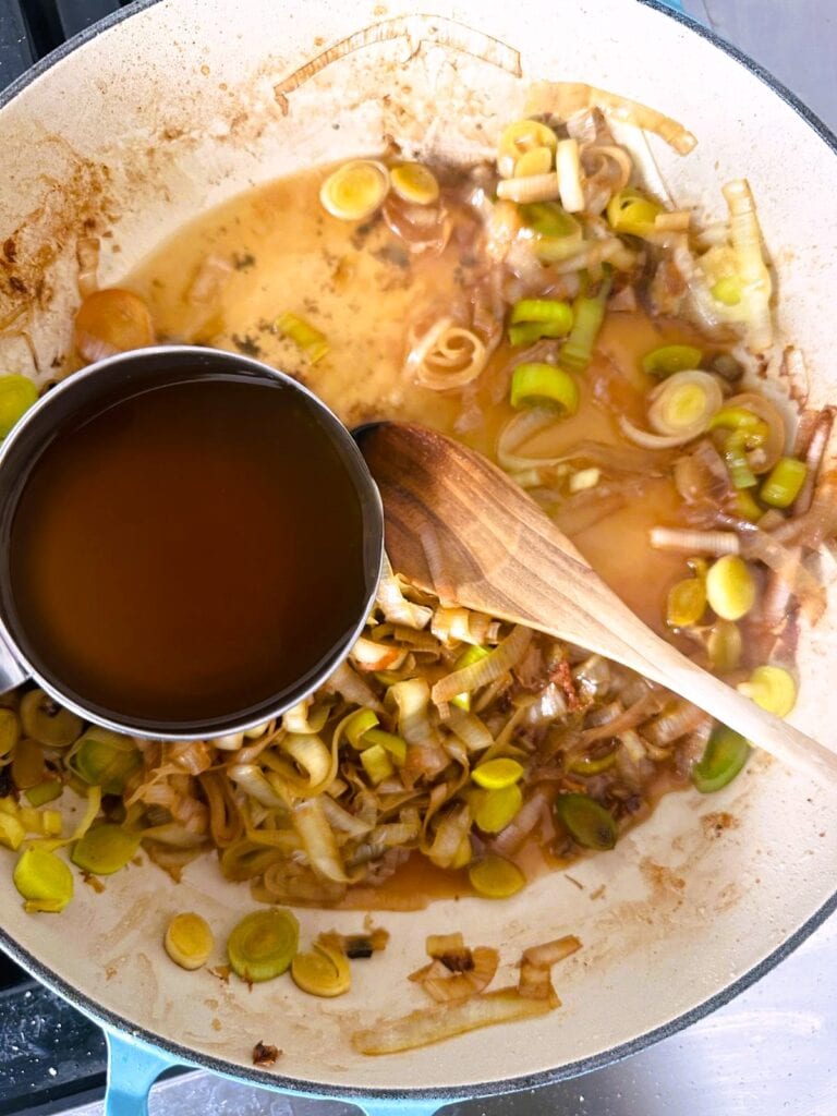 A large skillet of sauteed leeks with white wine being added to deglaze the pan.