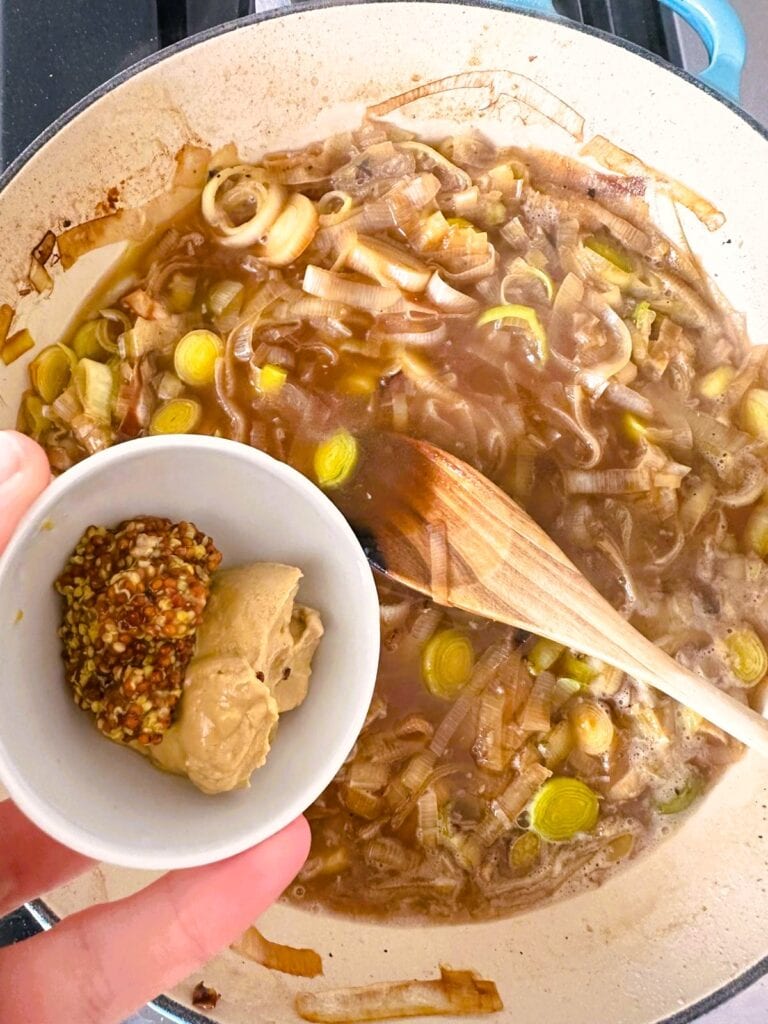 A large skillet of sauteed leeks in a stock and wine based sauce with wholegrain and Dijon mustard being added.