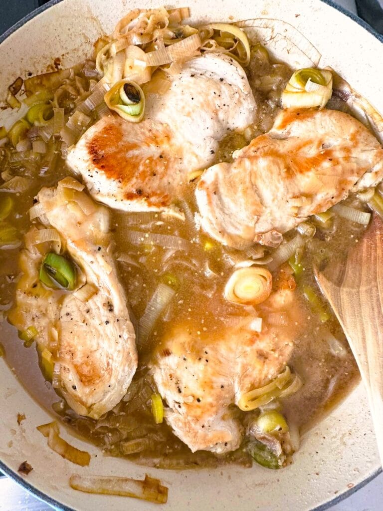 A large skillet of cooked chicken breasts with leeks and vegetable stock.
