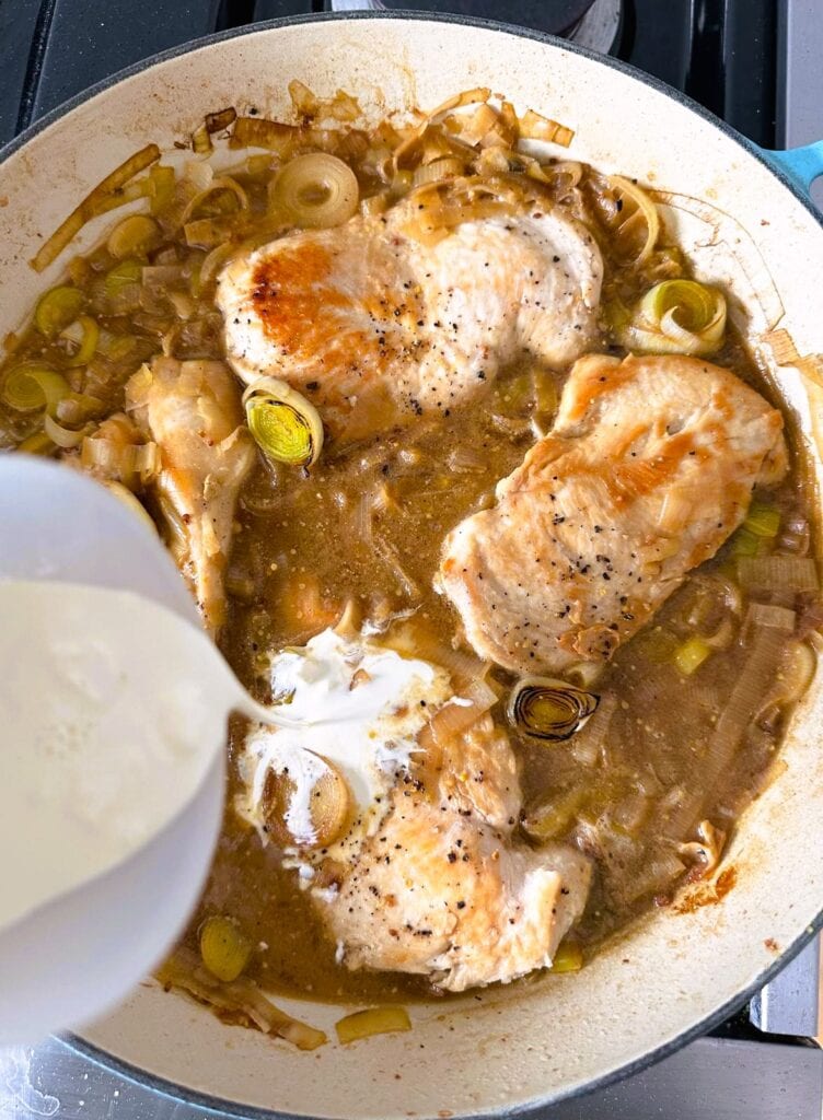 A large skillet of cooked chicken breasts and leeks in a stock based sauce with heavy cream being added.