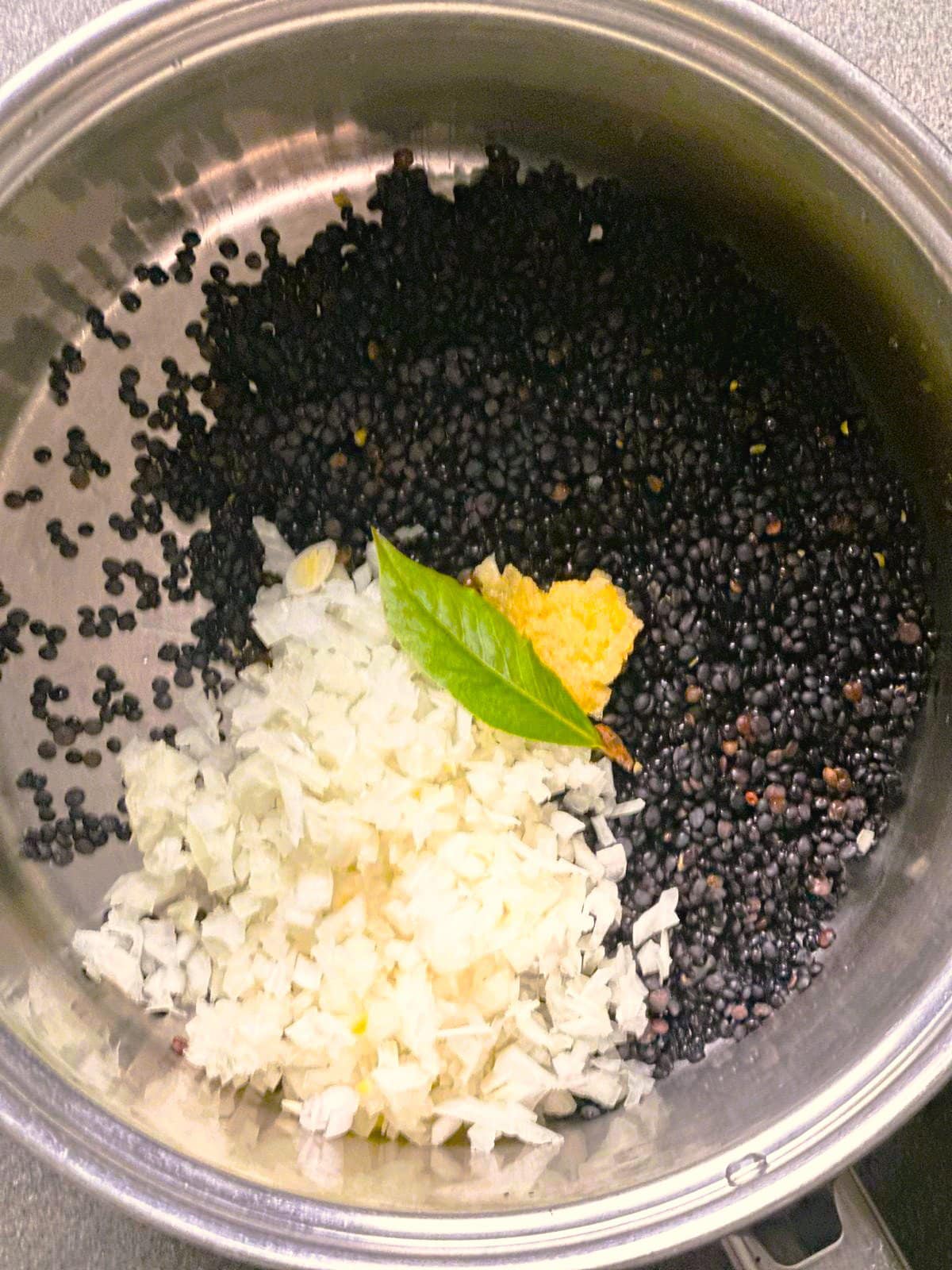 A large pot filled with dry black lentils, chopped onion and garlic and a bay leaf.
