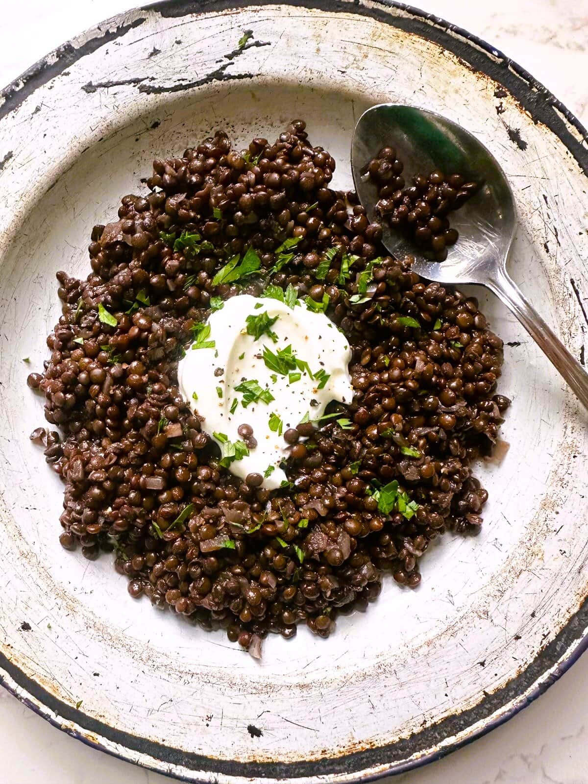 A bowl of black lentils (beluga lentils) topped with a spoon of thick Greek yogurt and a garnish of freshly chopped parsley.