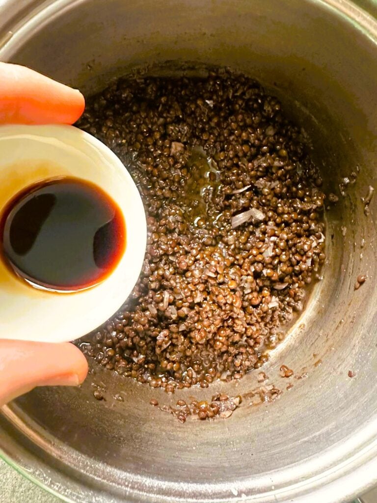 A large saucepan of cooked black lentils with balsamic vinegar being added.