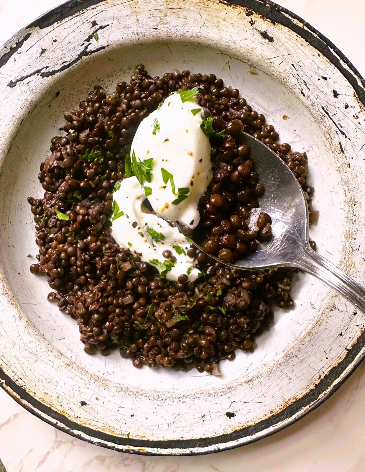 A bowl of black lentils (beluga lentils) topped with a spoon of thick Greek yogurt and a garnish of freshly chopped parsley.