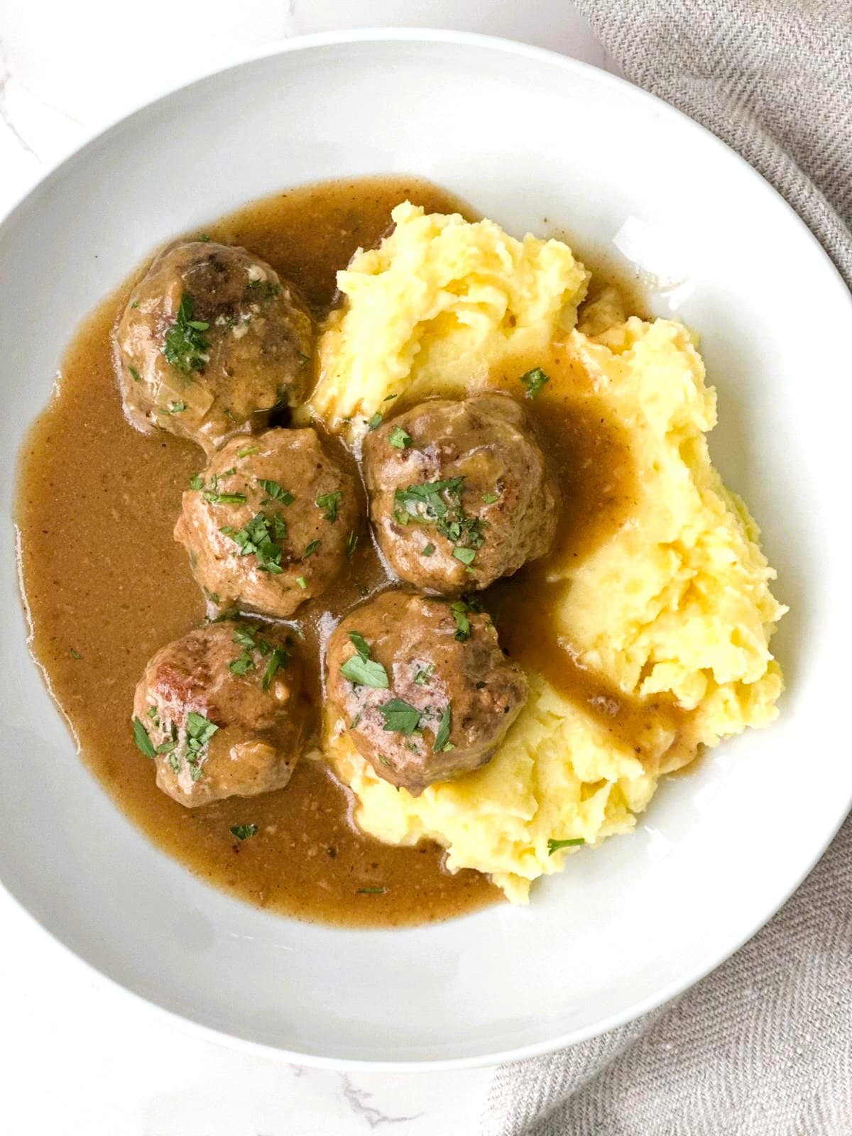A bowl of mashed potatoes topped with gluten-free meatballs in gravy, garnished with fresh parsley.