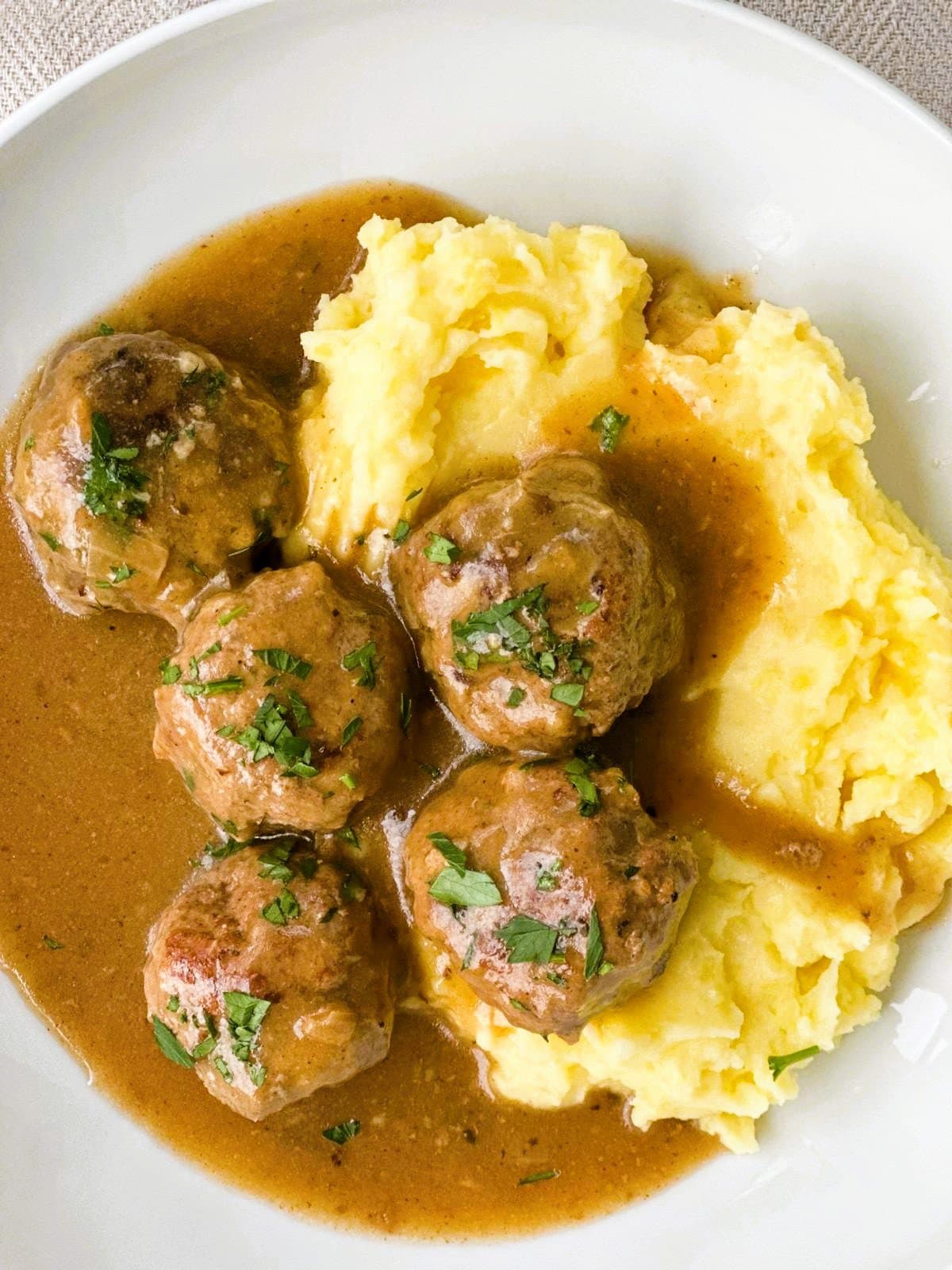 A bowl of mashed potatoes topped with gluten-free meatballs in gravy, garnished with fresh parsley.