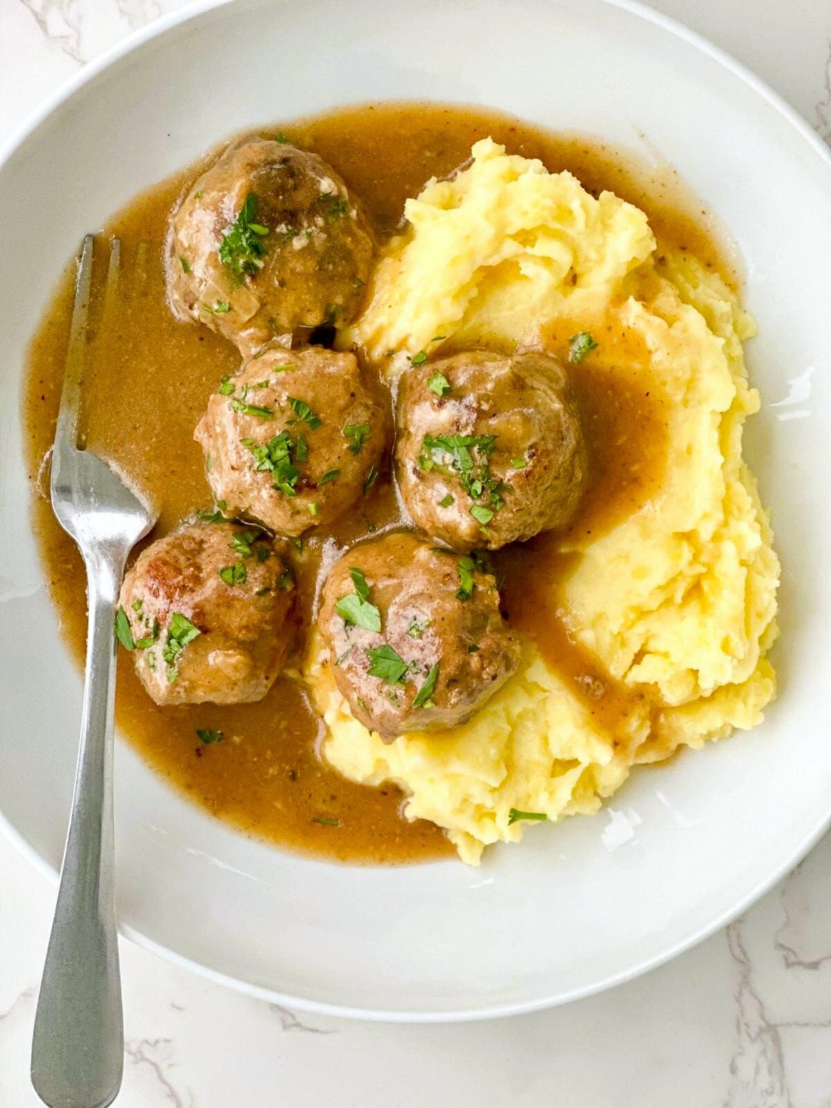 A bowl of mashed potatoes topped with gluten-free meatballs in gravy, garnished with fresh parsley.