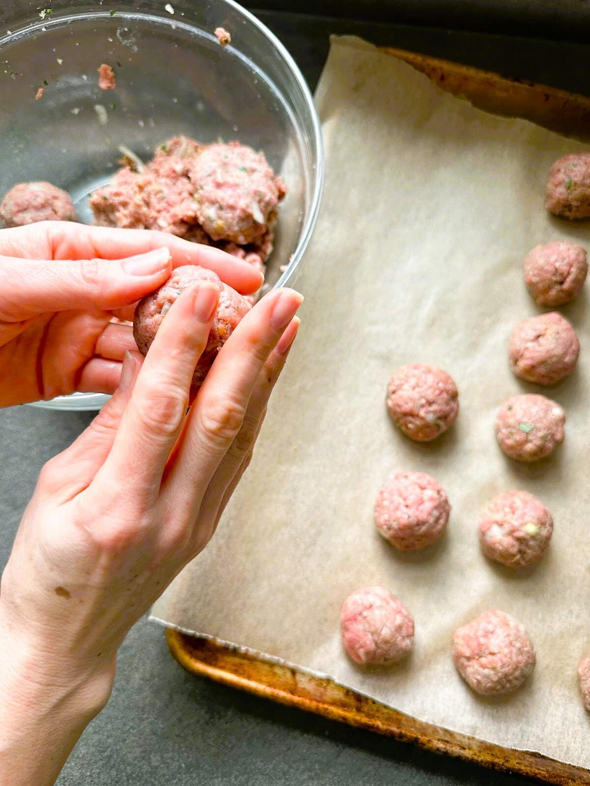 Image of a pair of hands rolling meatball mixture into even-sized balls with a line baking tray of meatballs set alongside.
