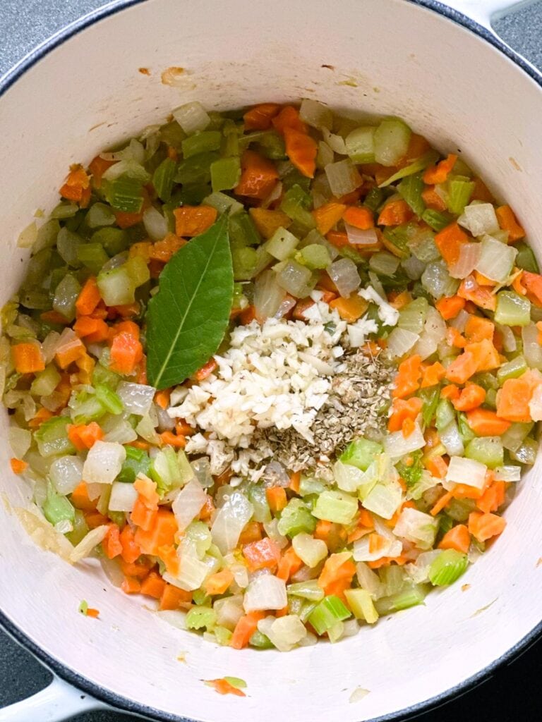 A Dutch oven with diced and sauteed onion, celery and carrot, with added freshly crush garlic, bay leaves and dried herbs.