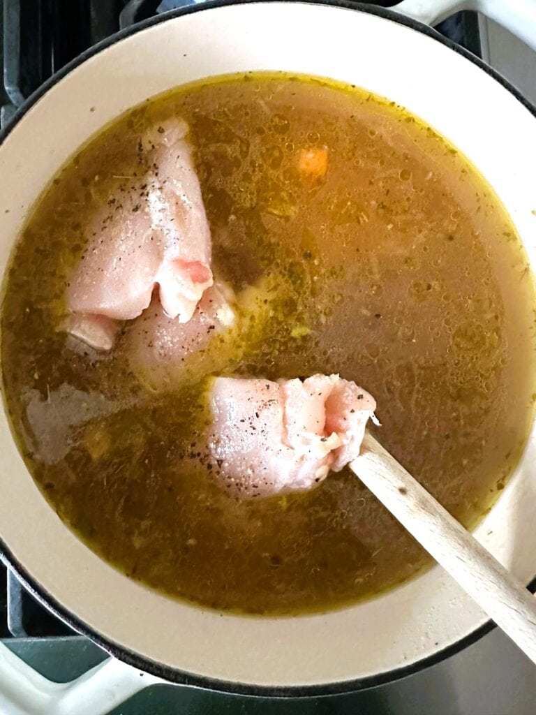 A Dutch Oven filled with vegetables and chicken stock with boneless, skinless chicken thighs being added.