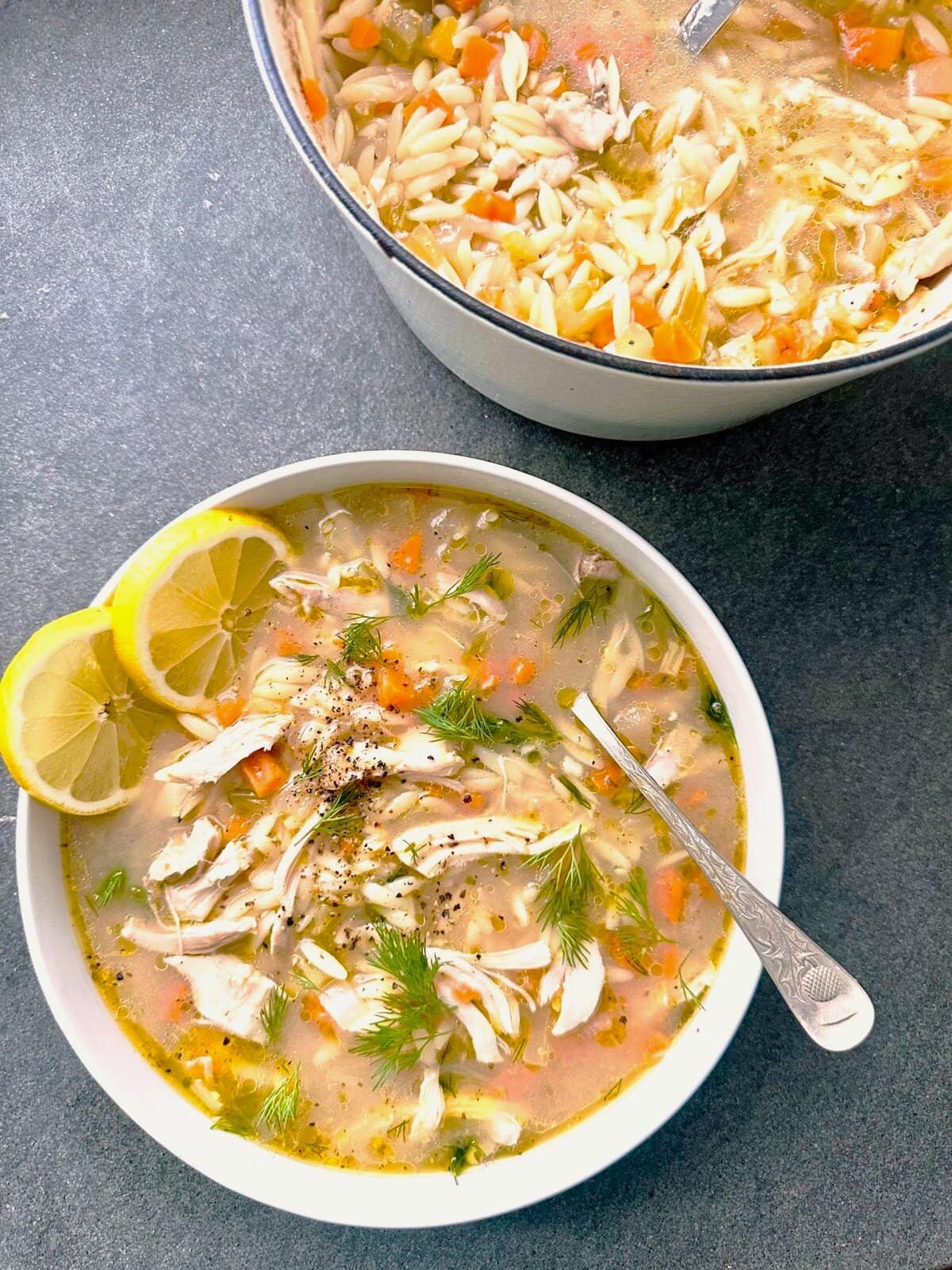 A bowl of lemon chicken orzo soup, garnished with fresh dill fronds and fresh lemon slices, with a Dutch oven of soup set alongside.