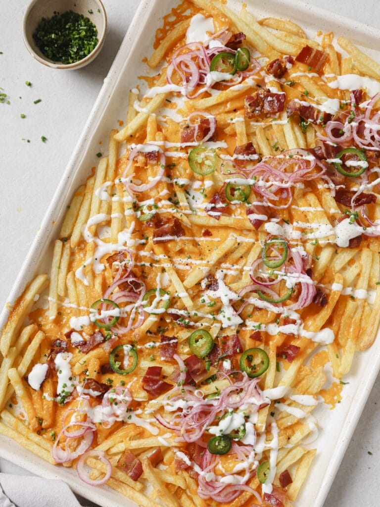 A lined baking sheet topped with golden French fries, crispy bacon pieces, melted cheese, pickled red onions and jalapenos, with a Greek yogurt drizzle and garnished with chopped chives.