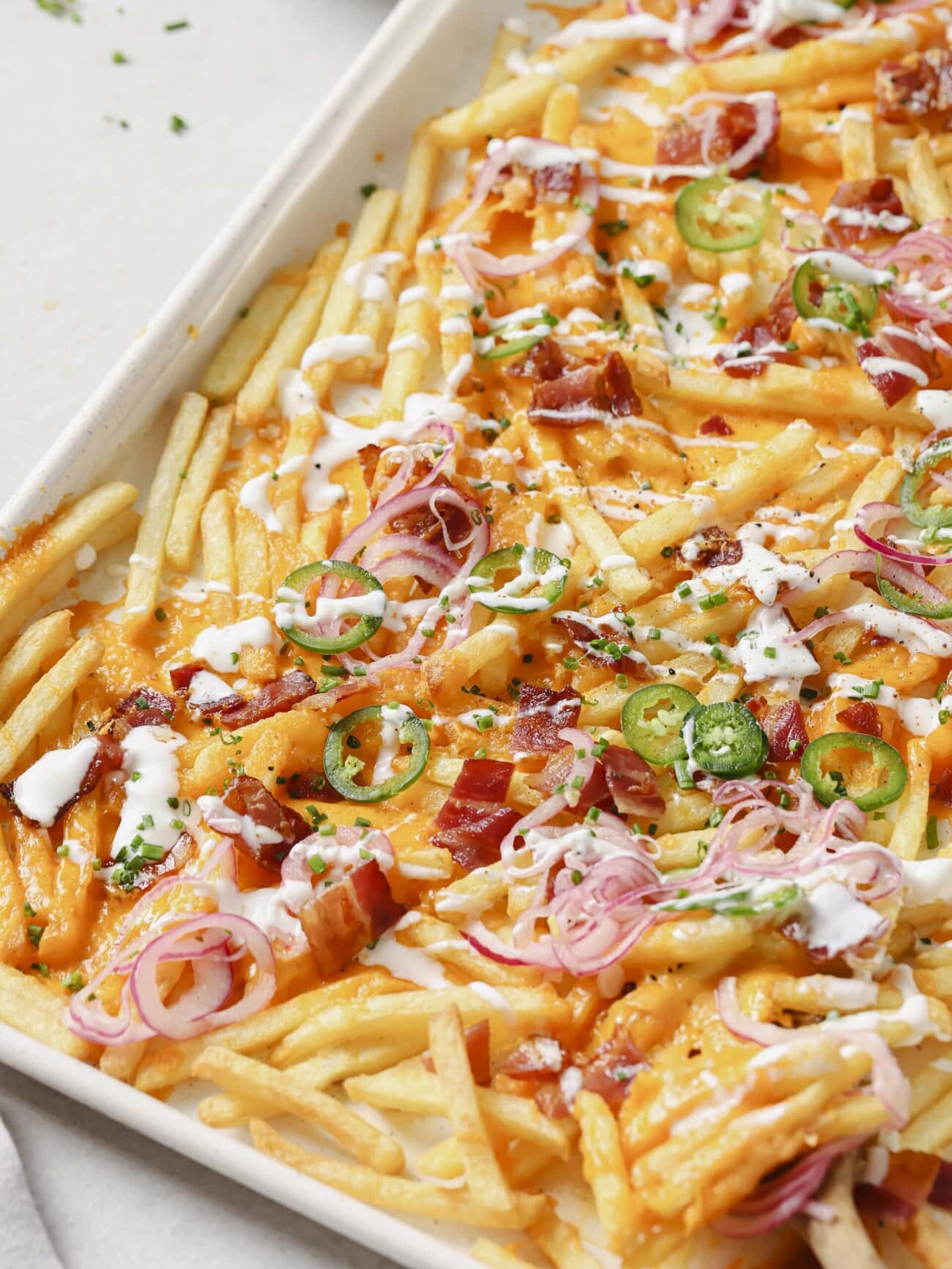 A lined baking sheet topped with golden French fries, crispy bacon pieces, melted cheese, pickled red onions and jalapenos, with a Greek yogurt drizzle and garnished with chopped chives.