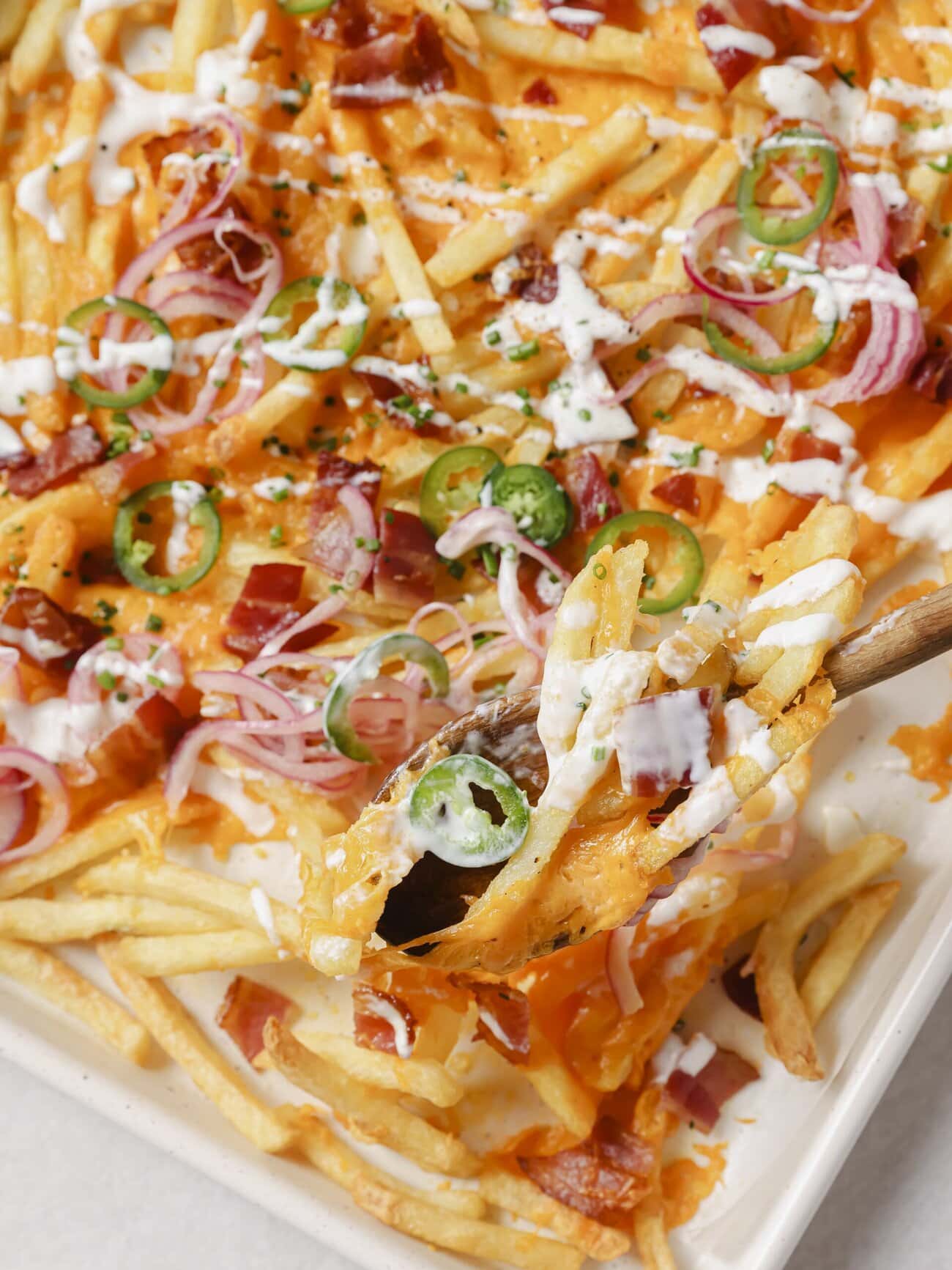 A lined baking sheet topped with golden French fries, crispy bacon pieces, melted cheese, pickled red onions and jalapenos, with a Greek yogurt drizzle and garnished with chopped chives.