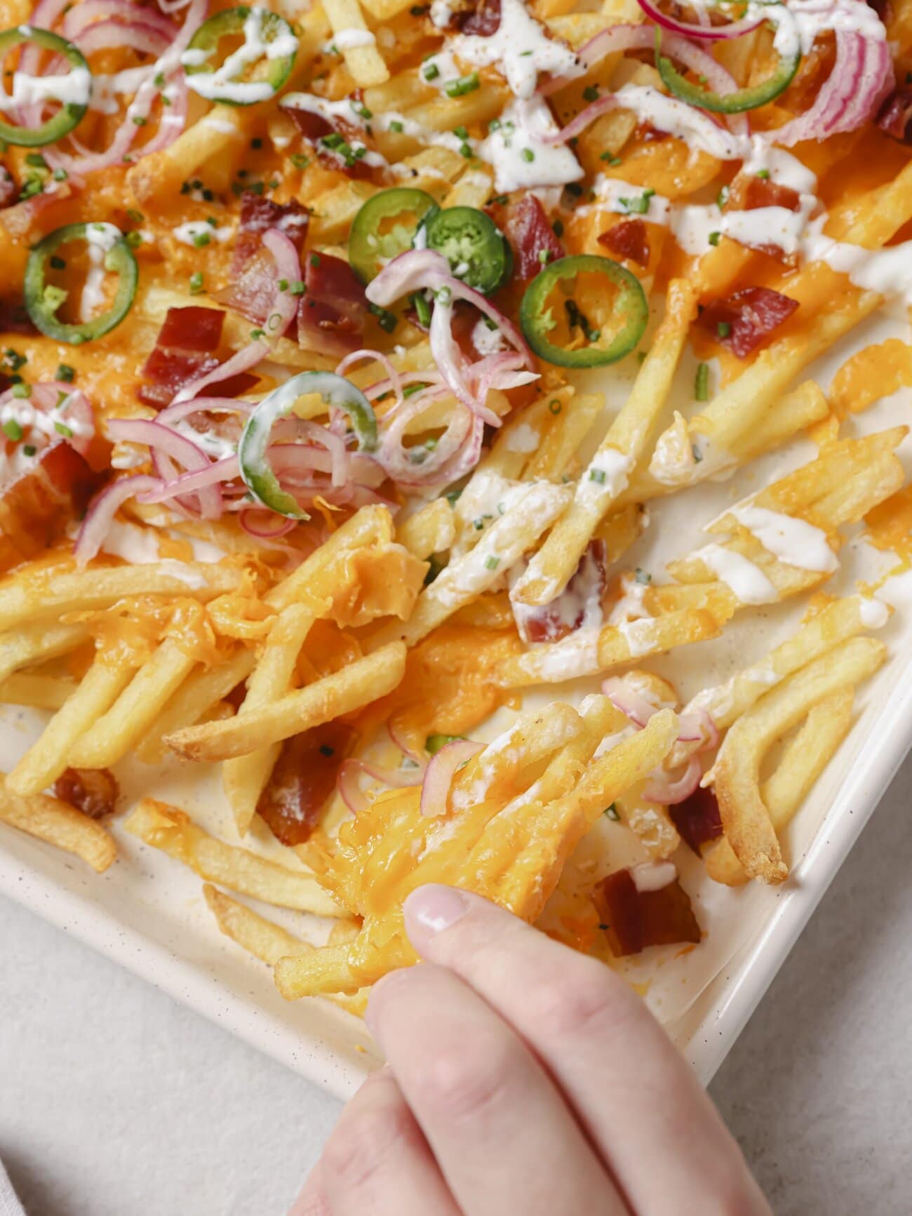 Close up image of golden French fries, crispy bacon pieces, melted cheese, pickled red onions and jalapenos, with a Greek yogurt drizzle and garnished with chopped chives.