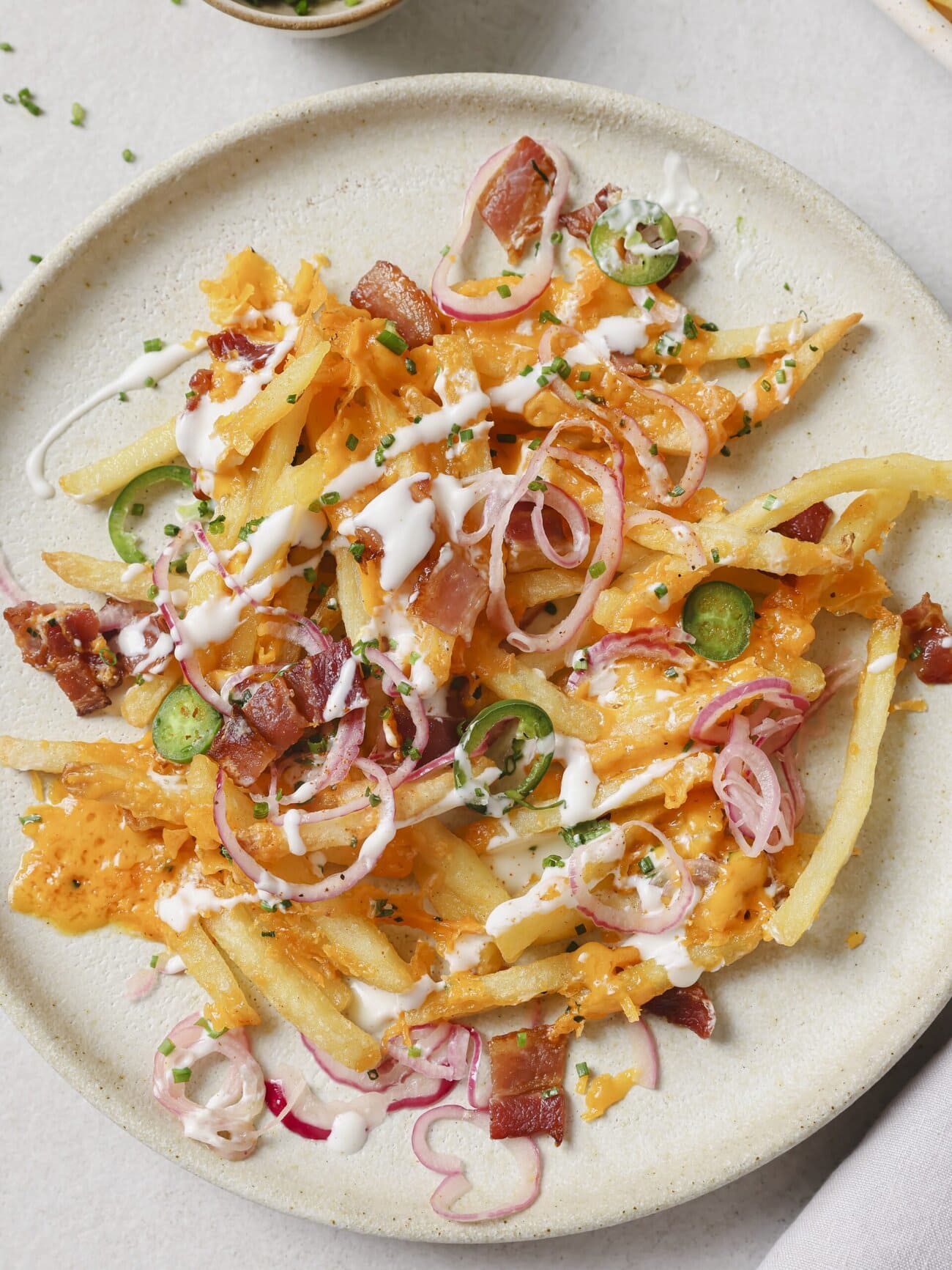 Aplate topped with golden French fries, crispy bacon pieces, melted cheese, pickled red onions and jalapenos, with a Greek yogurt drizzle and garnished with chopped chives.