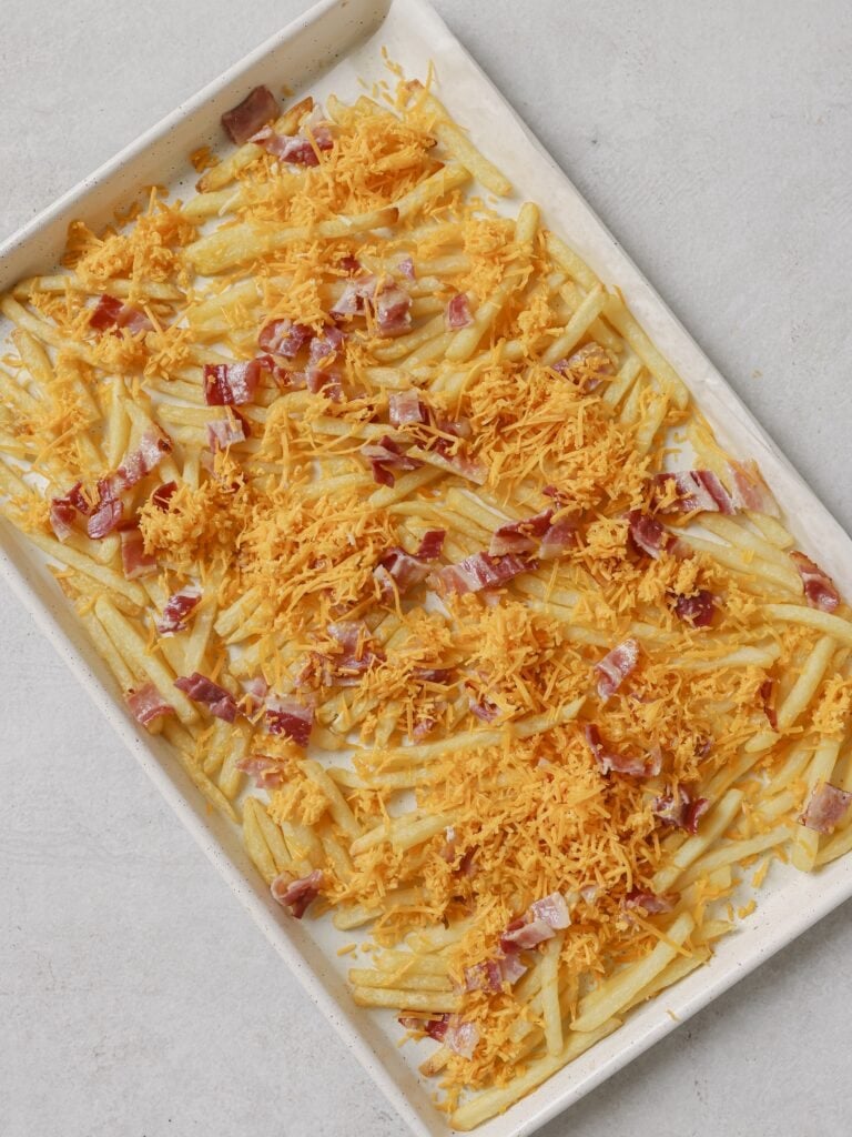 A lined baking sheet topped with cooked French fries, topped with cooked bacon pieces and shredded cheddar cheese.
