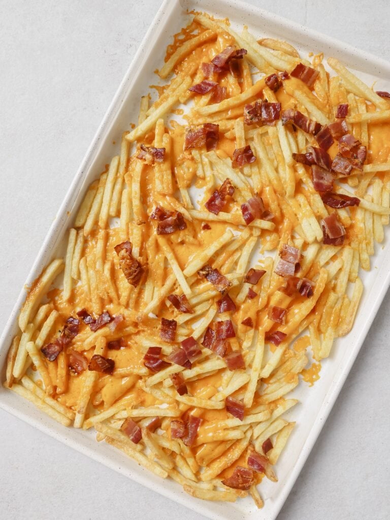 A lined baking sheet topped with cooked French fries, bacon pieces and melted cheddar cheese.