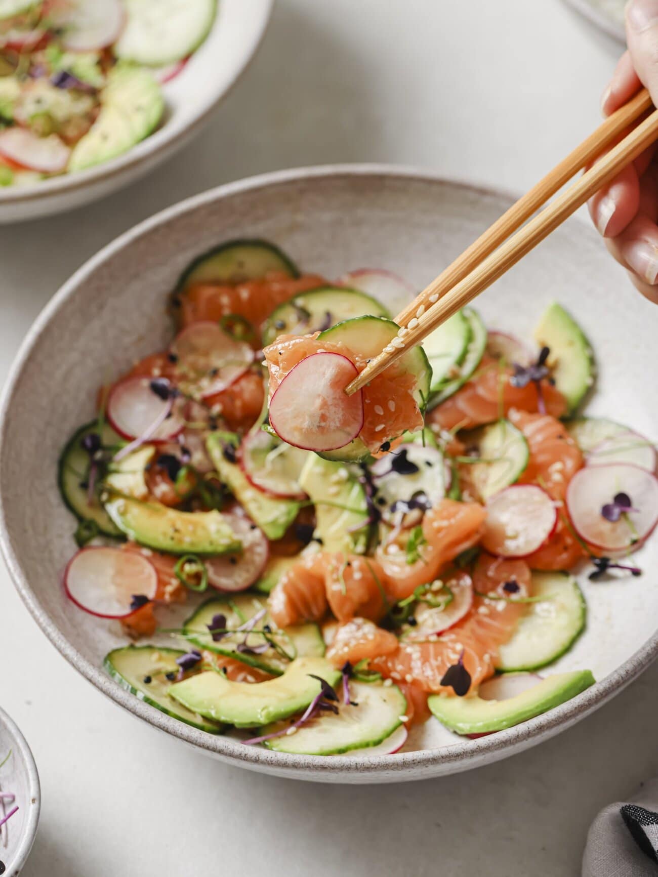 Salmon crudo bowl, comprising thin slices of sushi-grade salmon, with cucumber, avocado, radish and red chili, in a soy-lime honey dressing and garnished with microgreens and sesame seeds. With a pair of chopsticks holding some of the crudo.