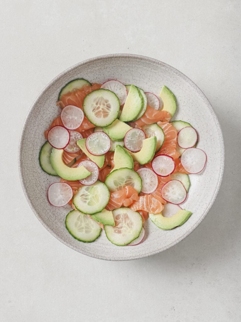 A bowl of thin slices of sushi-grade salmon, with cucumber, avocado, and radish.