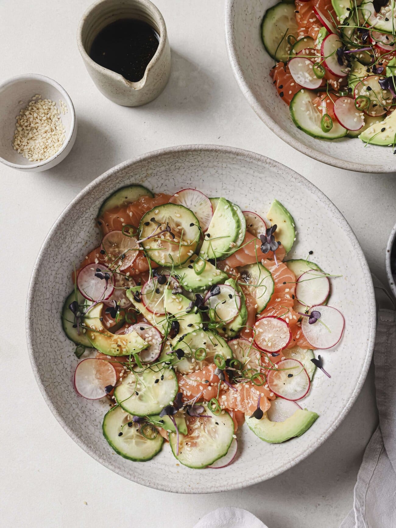 Salmon crudo bowl, comprising thin slices of sushi-grade salmon, with cucumber, avocado, radish and red chili, in a soy-lime honey dressing and garnished with microgreens and sesame seeds.