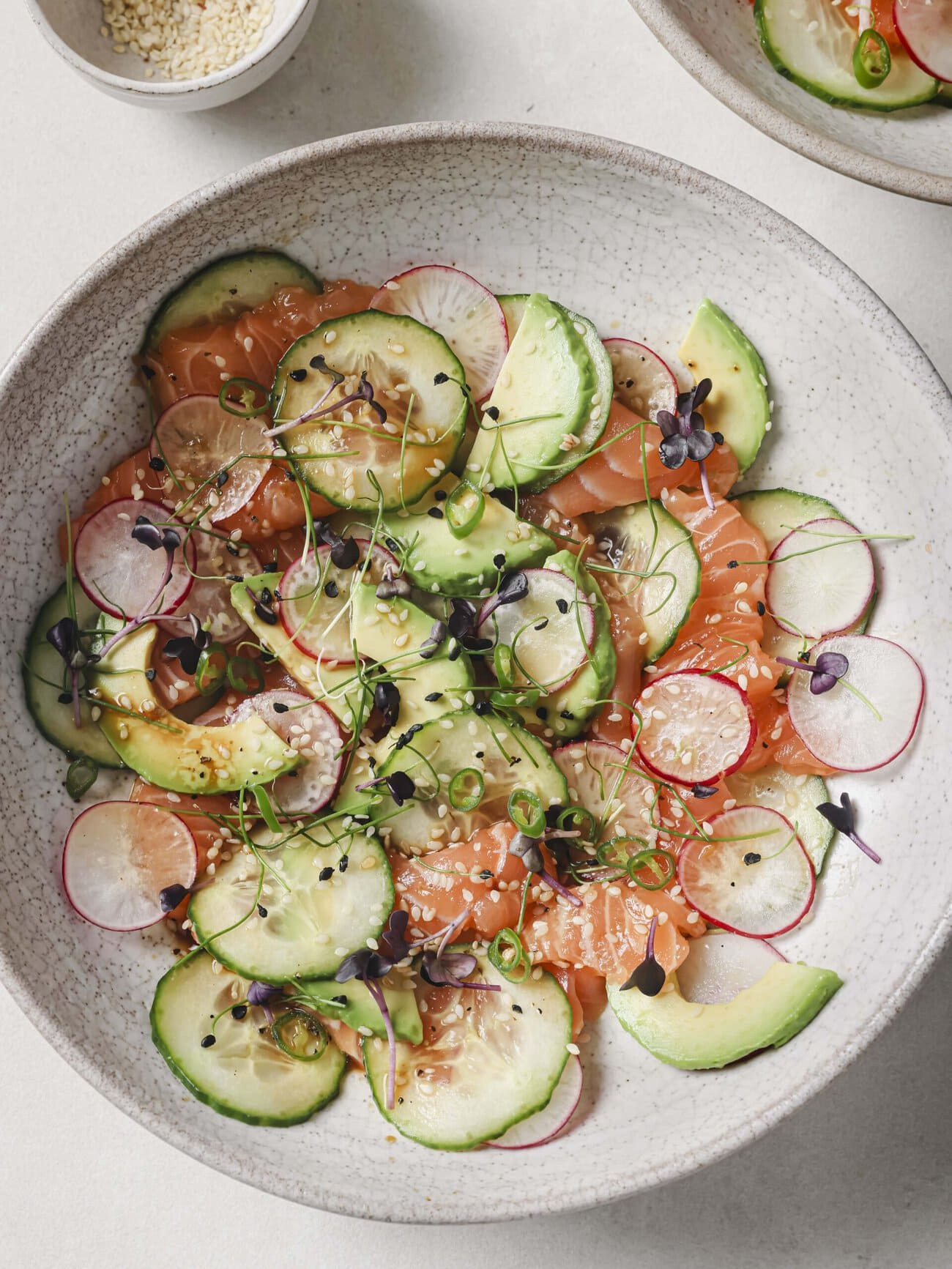 Salmon crudo bowl, comprising thin slices of sushi-grade salmon, with cucumber, avocado, radish and red chili, in a soy-lime honey dressing and garnished with microgreens and sesame seeds.