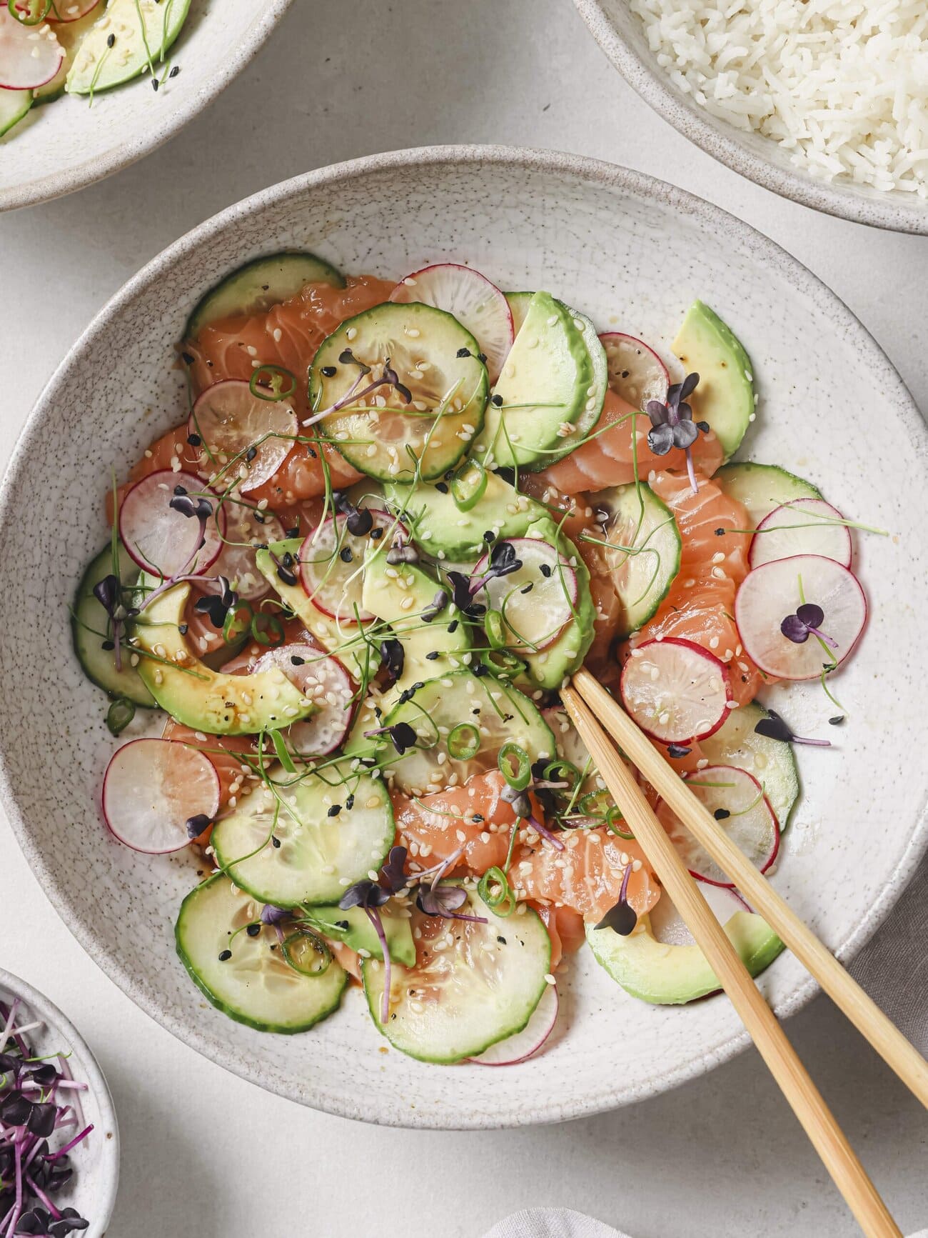 Salmon crudo bowl, comprising thin slices of sushi-grade salmon, with cucumber, avocado, radish and red chili, in a soy-lime honey dressing and garnished with microgreens and sesame seeds.