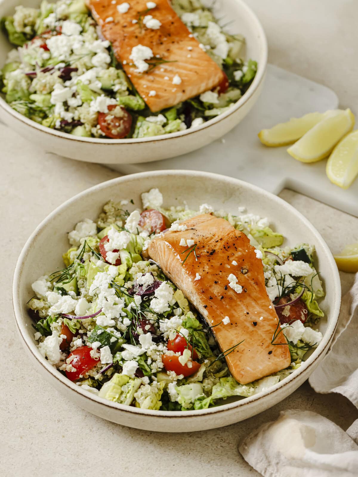 Two bowls of vibrant Greek Salmon with quinoa topped with fresh vegetables, feta, olives, and a creamy Green Goddess dressing.