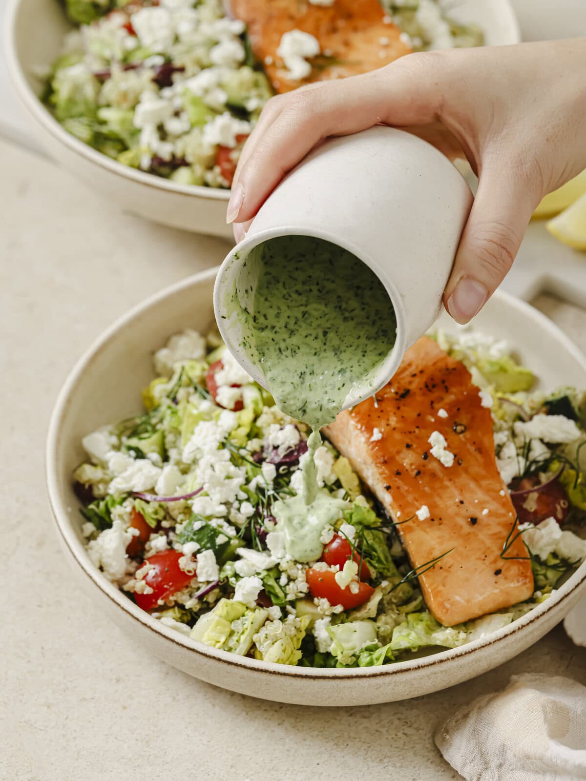 Two bowls of vibrant Greek Salmon with quinoa topped with fresh vegetables, feta, olives, and a creamy Green Goddess dressing being poured over.