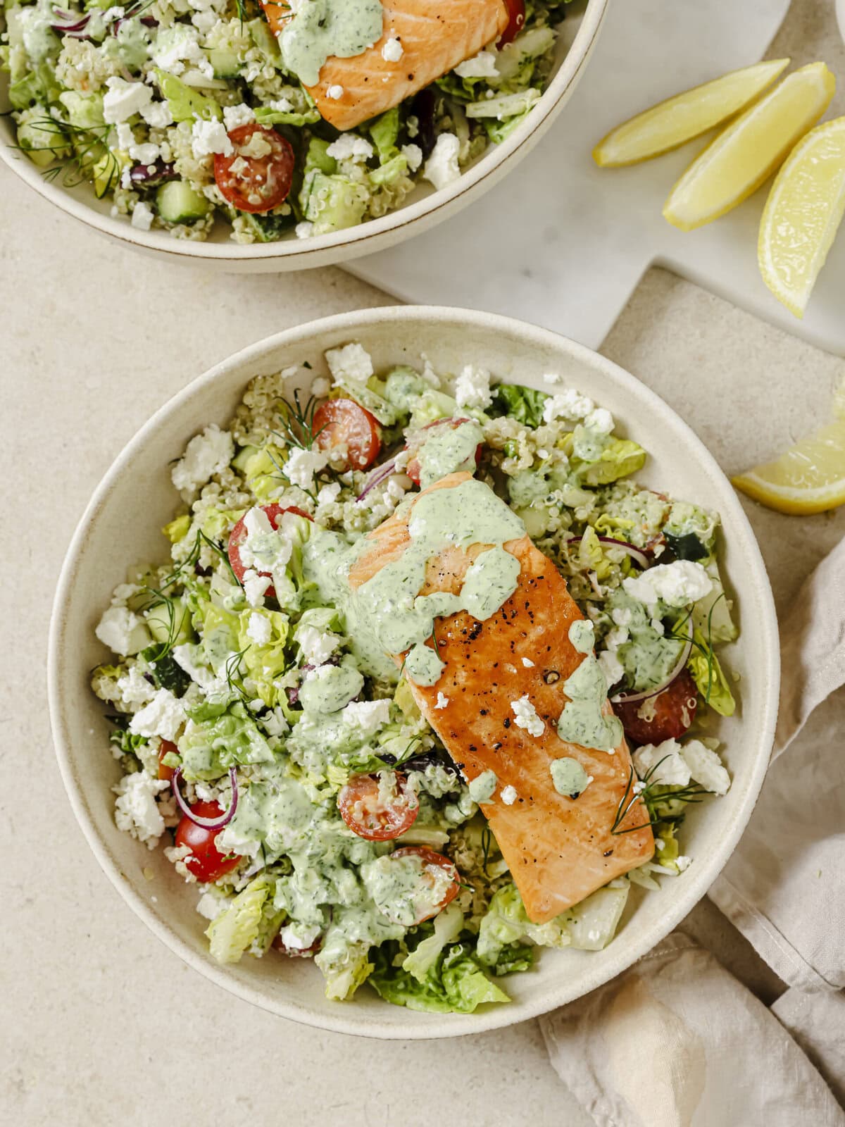 Two bowls of vibrant Greek Salmon with quinoa topped with fresh vegetables, feta, olives, and a creamy Green Goddess dressing.