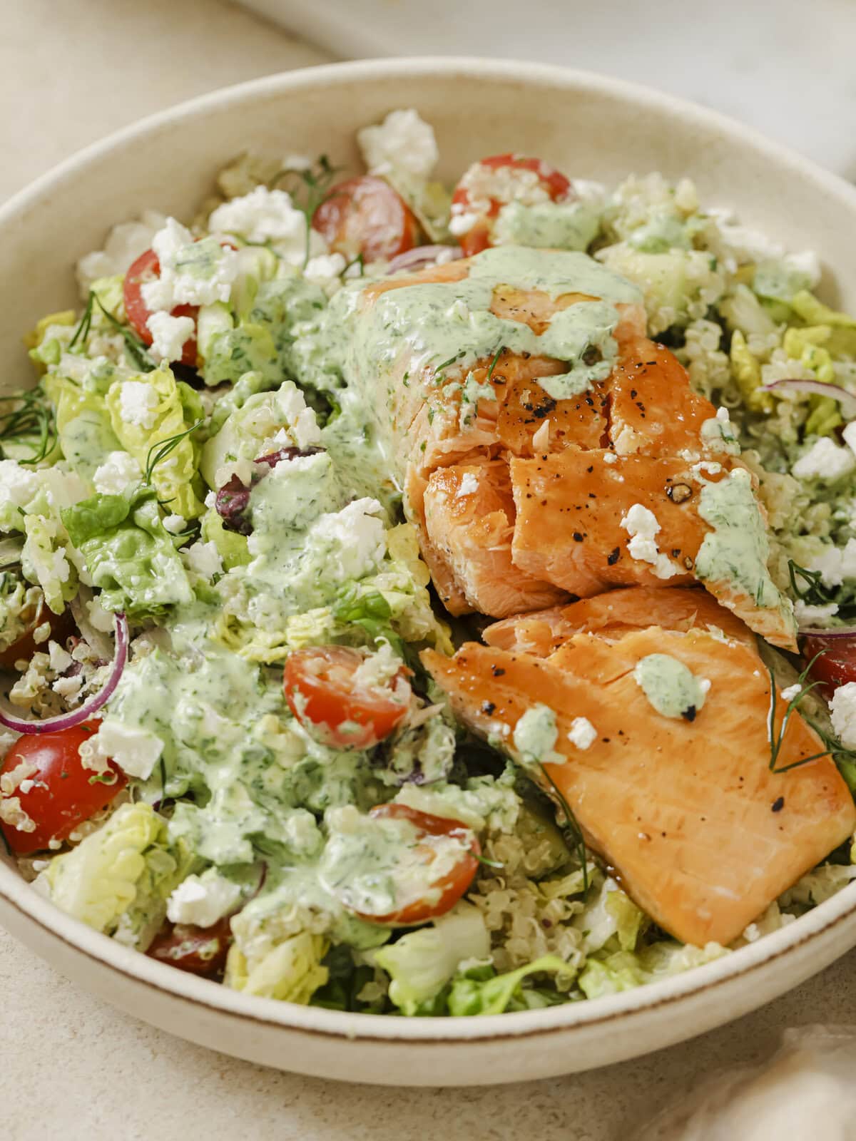 Close up image of vibrant Greek Salmon bowl with quinoa topped with fresh vegetables, feta, olives, and a creamy Green Goddess dressing.