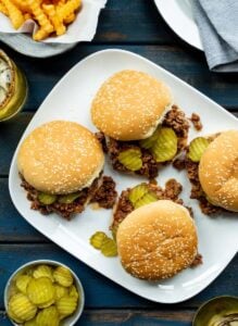 A white plate topped with 4 sloppy joe burger rolls, filled with ground beef and sliced pickles.