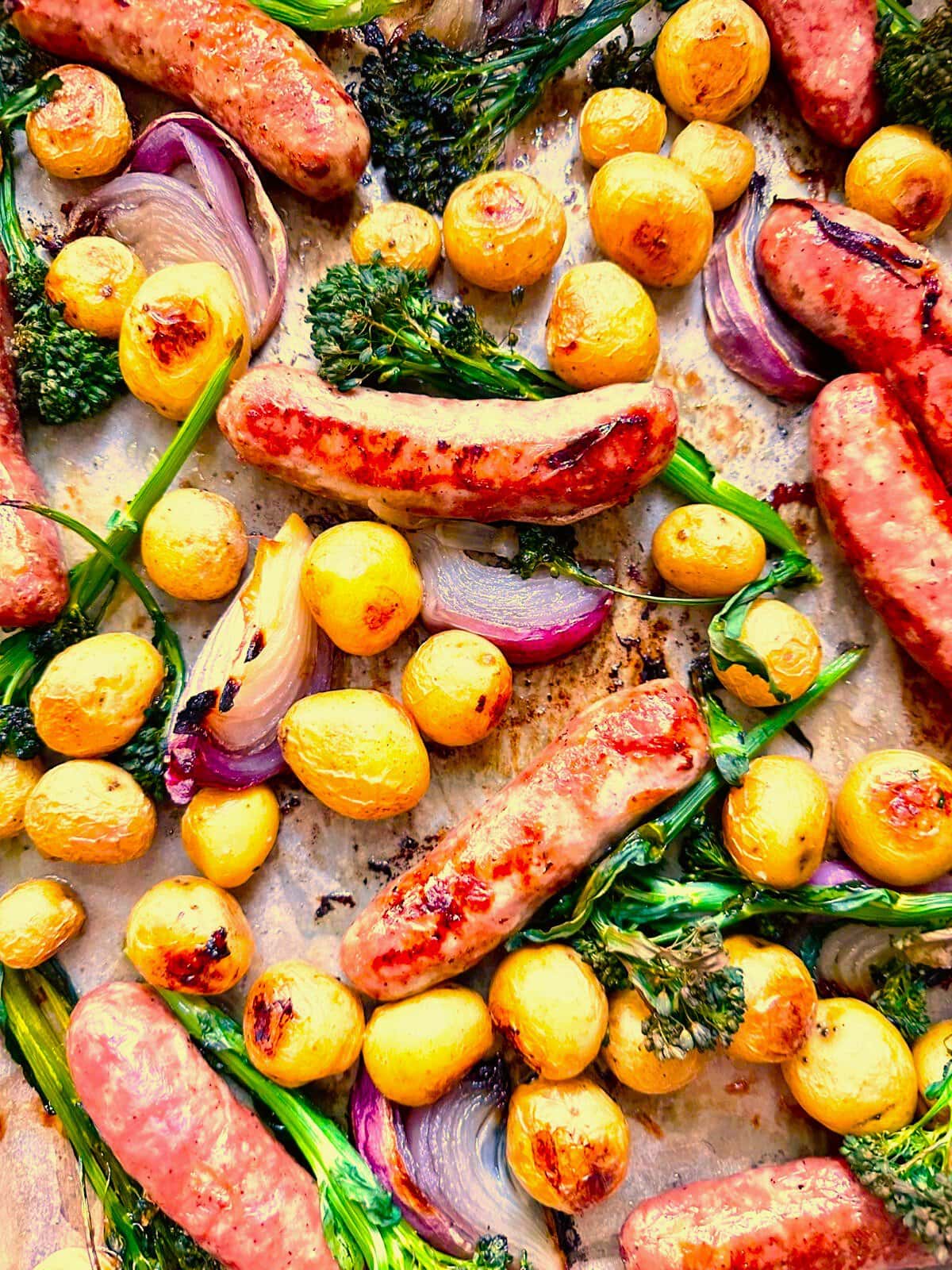 Close up image of sheet pan topped with roasted sausages, baby potatoes, broccolini, and red onion, served with a Greek yogurt and lemon dressing.