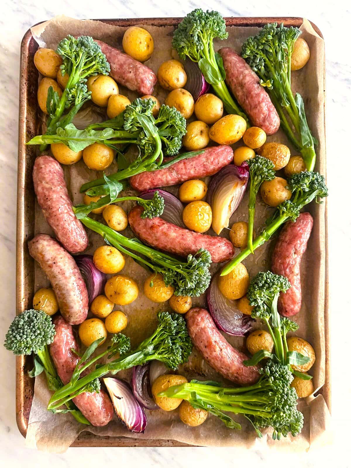 Sheet pan topped with roasted sausages, baby potatoes, broccolini, and red onion.