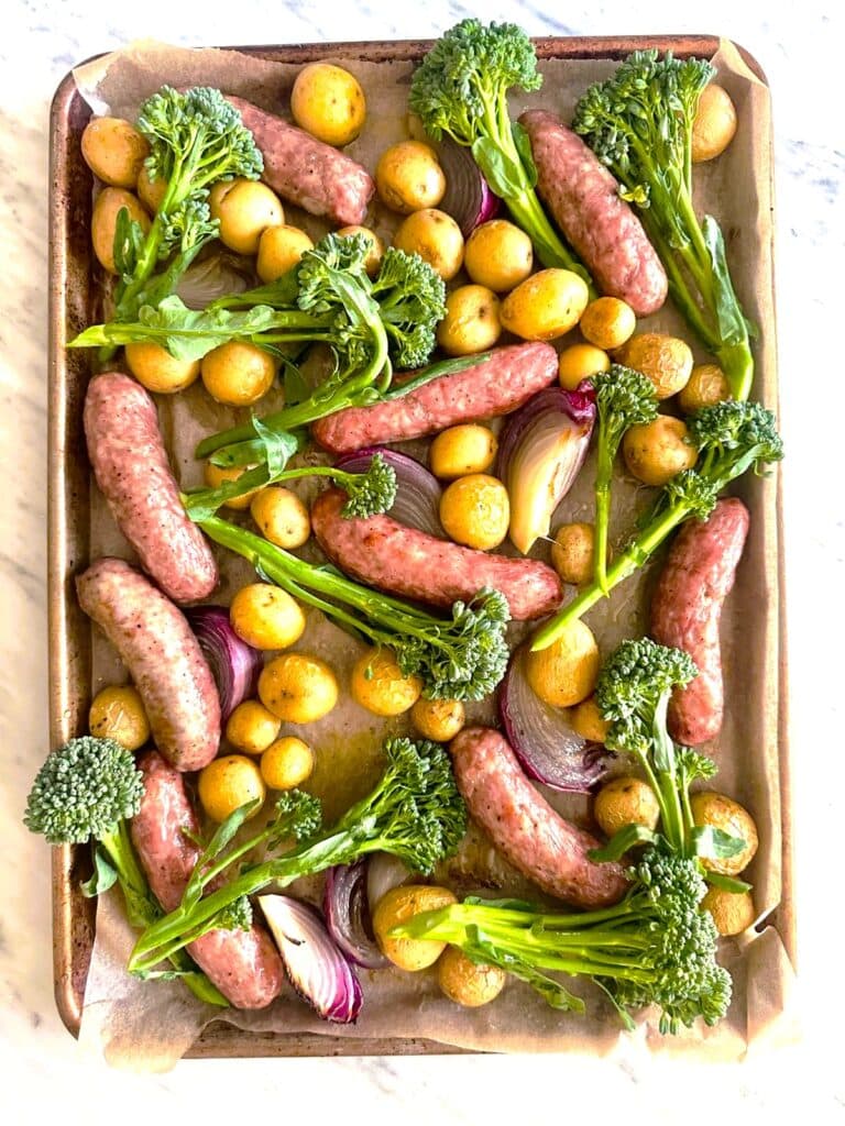 Paper lined sheet pan topped with part cooked Italian sausages, baby potatoes and sliced red onion, with added broccolini.