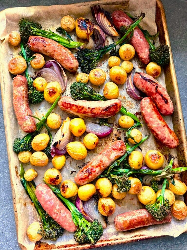 Sheet pan topped with roasted sausages, baby potatoes, broccolini, and red onion.