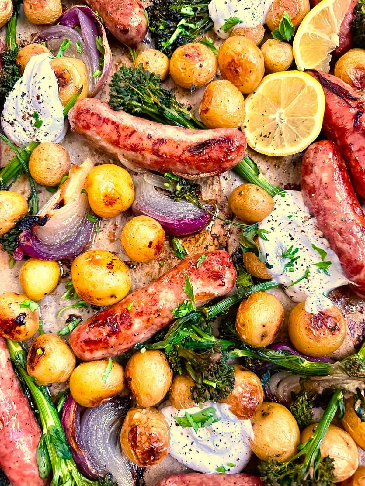 Close up image of a sheet pan topped with roasted sausages, baby potatoes, broccolini, and red onion, served with a Greek yogurt and lemon dressing.