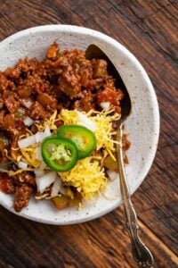 A white bowl of ground beef chilli topped with shredded cheese, diced onion and sliced jalapeno.